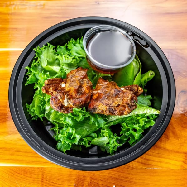 Grilled chicken pieces on a bed of fresh green lettuce with a side of dark sauce in a black bowl.