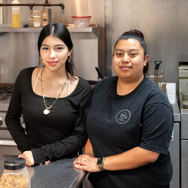 Two smiling Pokefit staff members in black shirts stand behind a counter in a commercial kitchen.