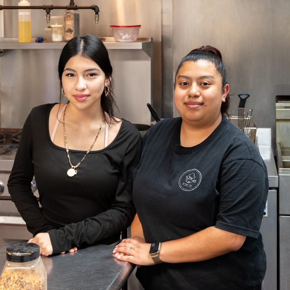 Two smiling Pokefit staff members in black shirts stand behind a counter in a commercial kitchen.