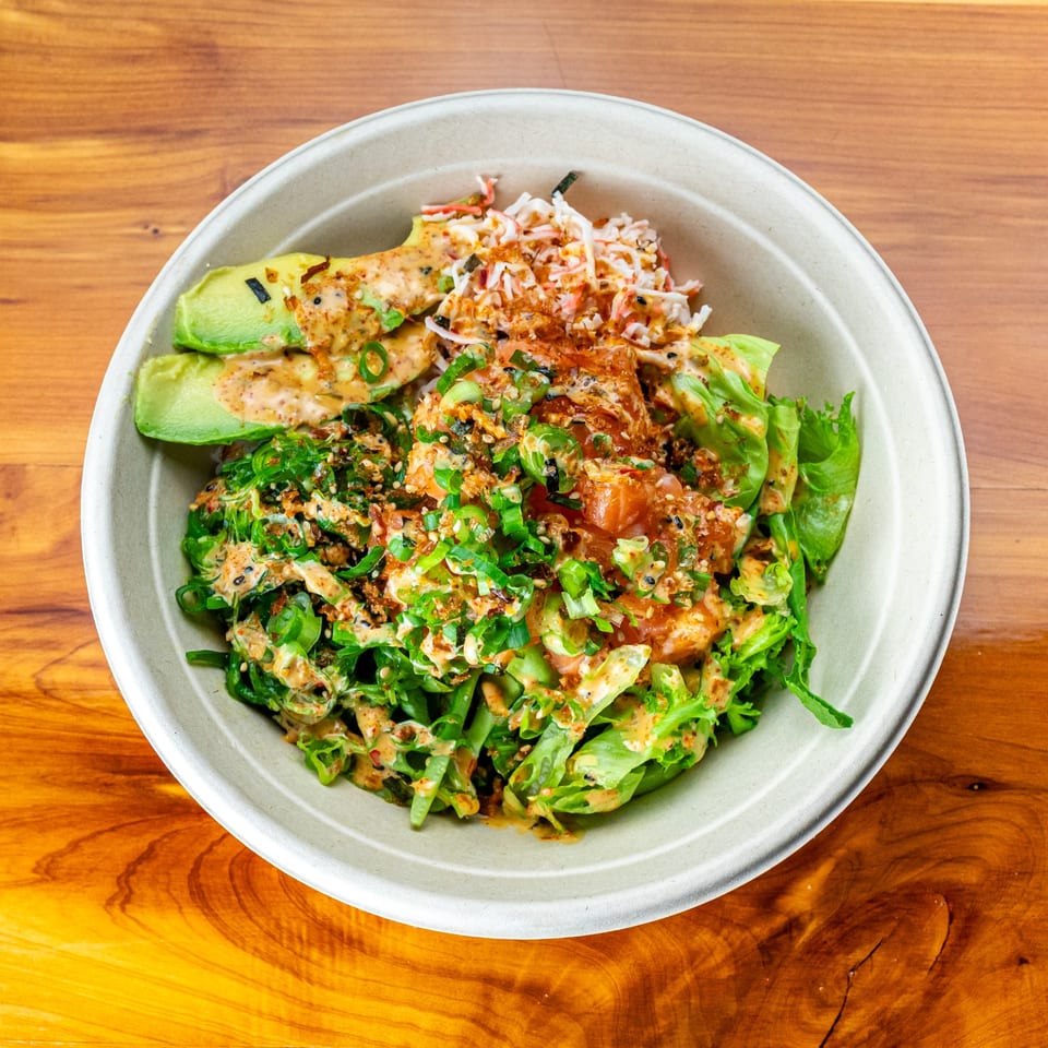 A vibrant Pokefit poke bowl with salmon, avocado, crab, greens, and sauce on a wooden table.