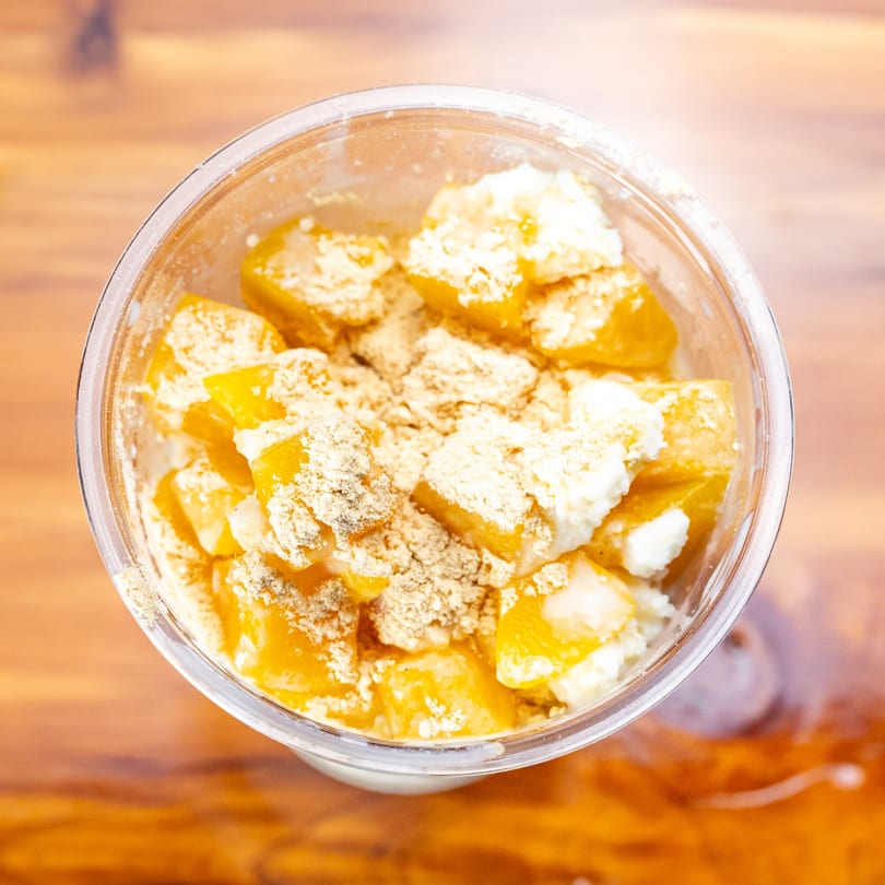 Overhead view of a clear cup filled with diced mango, white cream, and a brown powder.