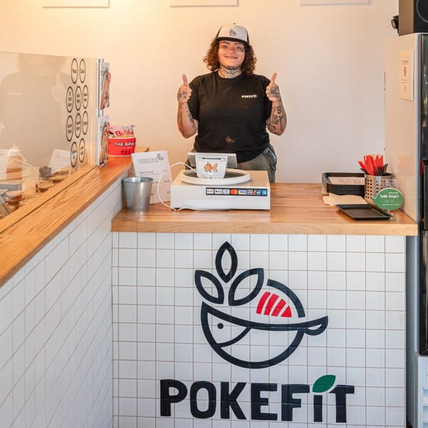 A Pokefit employee in a black shirt and cap gives two thumbs up behind the counter.