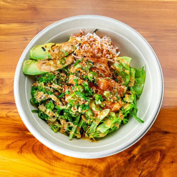 A Pokefit poke bowl with salmon, avocado, crab, lettuce, and green onions on a wooden table.