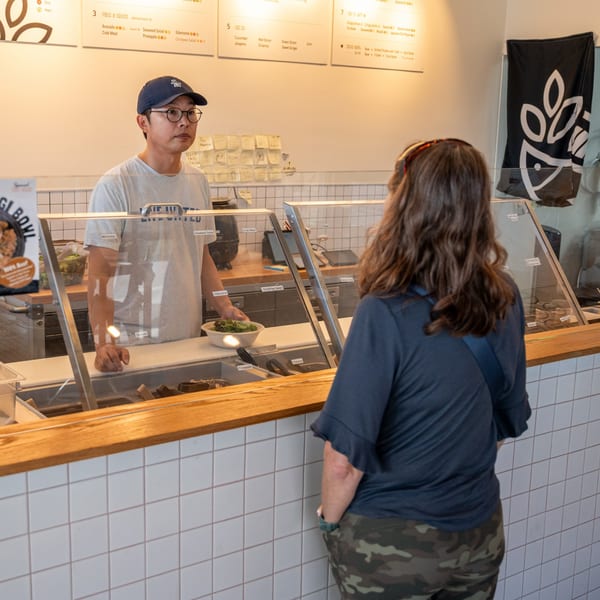 A Pokefit employee serves a customer at the counter, with menu boards visible in the background.