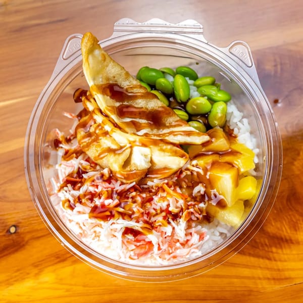 A Pokefit poke bowl with rice, crab, edamame, pineapple, and fried dumplings drizzled with sauce.
