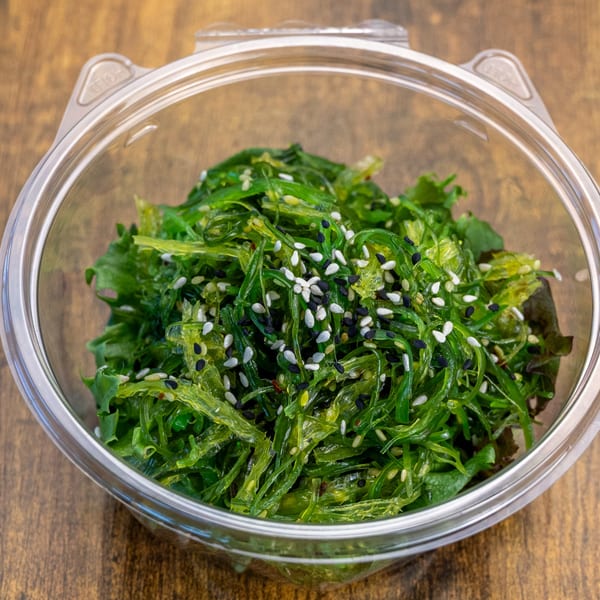 Pokefit's vibrant green seaweed salad, topped with white and black sesame seeds, in a clear plastic container.