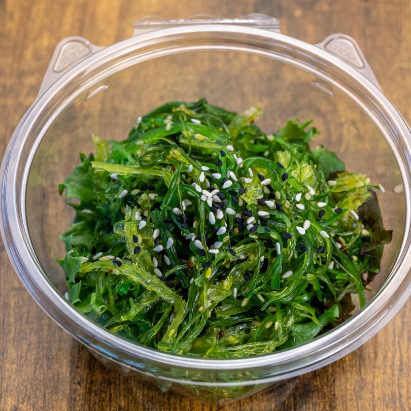Pokefit's vibrant green seaweed salad, topped with white and black sesame seeds, in a clear plastic container.