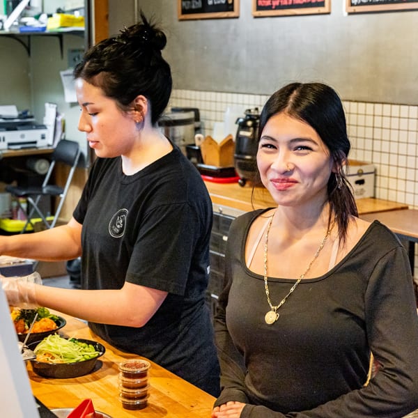 Two Pokefit employees, one preparing food and the other smiling at the camera.