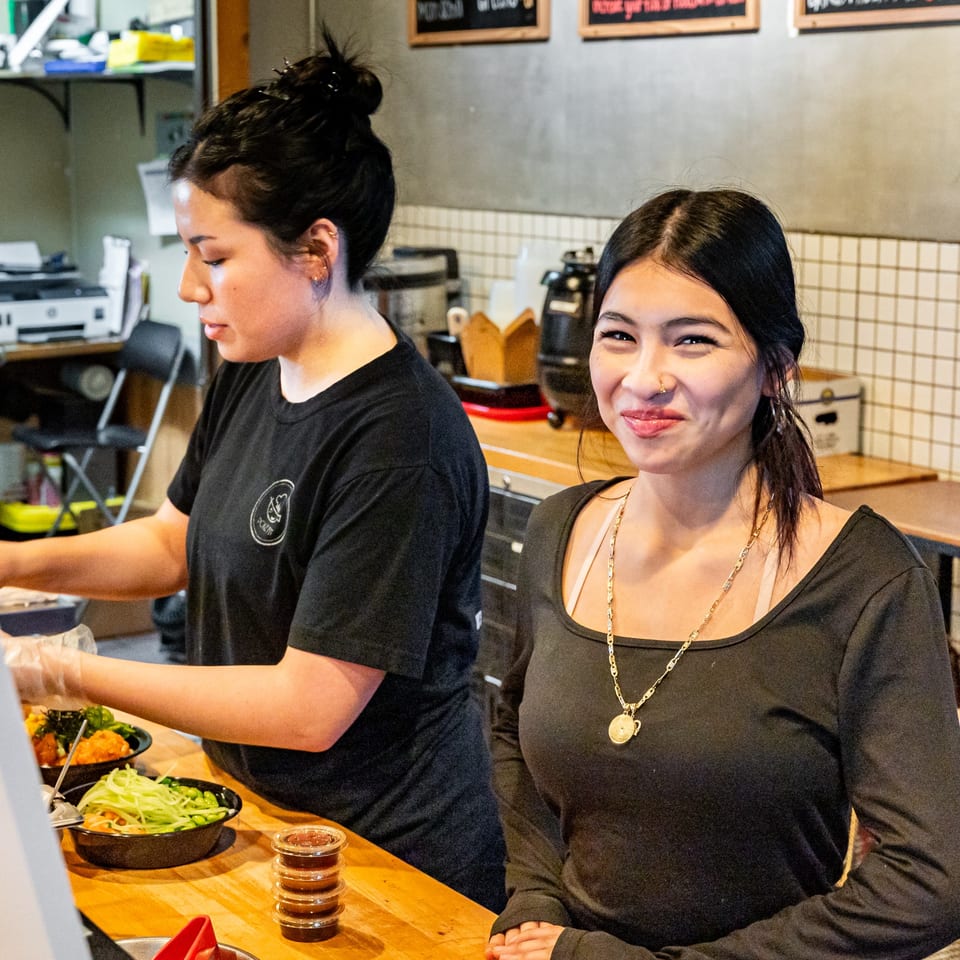 Two Pokefit employees, one preparing food and the other smiling at the camera.