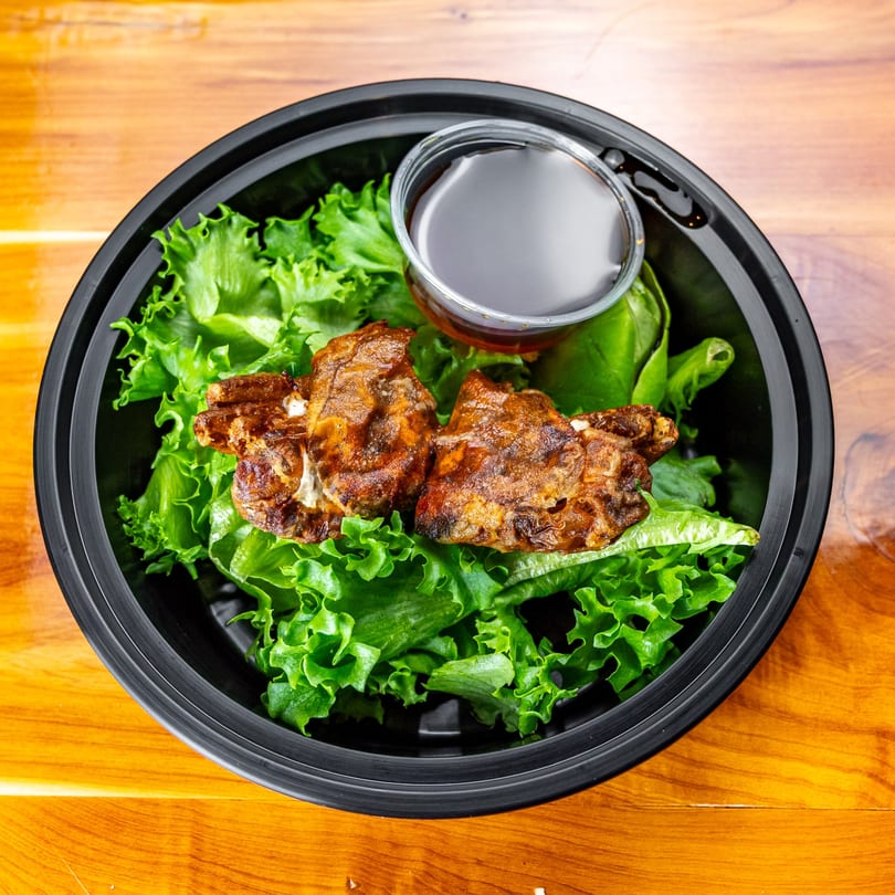 Grilled chicken pieces on a bed of fresh green lettuce with a side of sauce in a black bowl.
